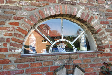 Halvrundt vindue med porcelænsfigurer i vindueskarmen og spejling af et rødt hus i baggrunden.