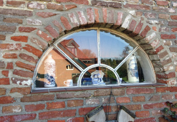 Ventana semicircular de ladrillo con figuras de cerámica y reflejo de una casa roja del exterior.