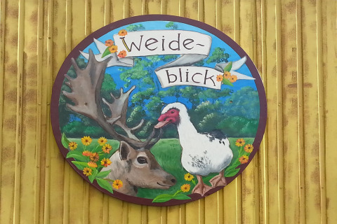 Insegna dipinta con renna, oca e fiori, testo: 'Weide-blick', su parete gialla a Pipowagen nei Paesi Bassi.