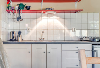 Moderne køkkenindretning med hvide skabe, røde hylder, flisebeklædt væg og køkkenudstyr i et feriehus.