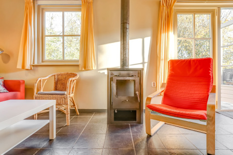 Living room with stove, red chair, wicker chair, and large windows at Holiday Home 5 Aarde Green Camping Lage Zwaluwe.