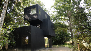 Resort Gulpen 8 modern black lodge, surrounded by tall trees, set next to a dirt path in the forest.
