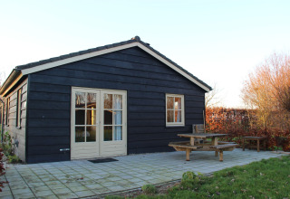 Sort trælodge med beige dør og vindue, terrasse med bænk, beliggende på Holiday Home 3 Aarde Green Camping Lage Zwaluwe, Holland.