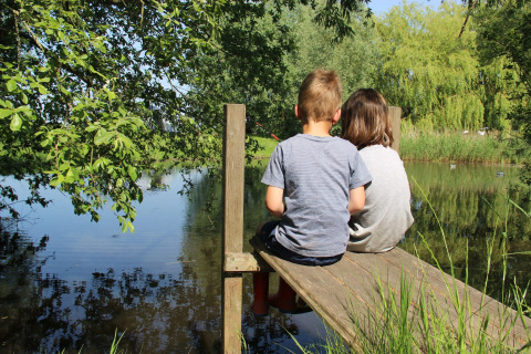 Two children sit on a wooden dock by a lake at Holiday Home 3 Aarde Green Camping Lage Zwaluwe in the Netherlands.