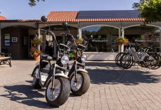 Dos scooters eléctricos y varias bicicletas frente al minimercado en Park Duc de Brabant 3, Países Bajos.
