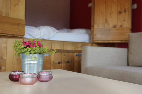 Gemütliches Zimmer mit Holzmöbeln, Bett, Sofa, Blumentopf und Teelichthaltern im Vordergrund.