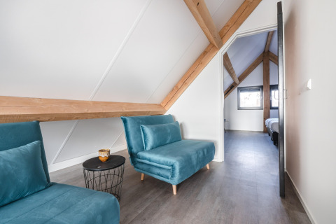 Moderne loftstue i Park de Kievit 22, Belgien, med blå stole, træbjælker og udsigt mod soveværelse.