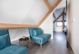 Moderne loftstue i Park de Kievit 22, Belgien, med blå stole, træbjælker og udsigt mod soveværelse.