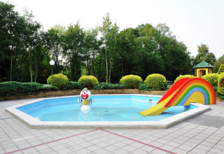 Piscina infantil en un chalet con una estatua de payaso y un tobogán arcoíris, rodeada de vegetación.