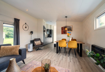 Moderne opholdsstue og spiseområde med gule stole, åbent køkken og store vinduer i Brabants Weelde 33-lodge.