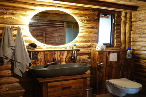 Rustikt badeværelse med trævægge, rund oplyst spejl, moderne håndvask og væghængt toilet i chalet.