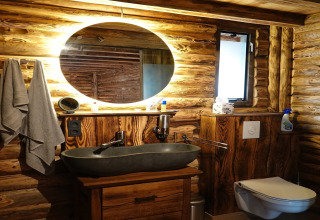 Rustikt badeværelse med trævægge, rund oplyst spejl, moderne håndvask og væghængt toilet i chalet.