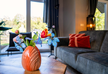 Stue i Holiday Park «Latour 4» med orange vase og blomster på bordet, sofa og store vinduer i baggrunden.