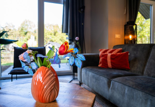 Holiday Park «Latour 4» lounge with an orange vase and flowers on a table, grey sofa, and large windows.
