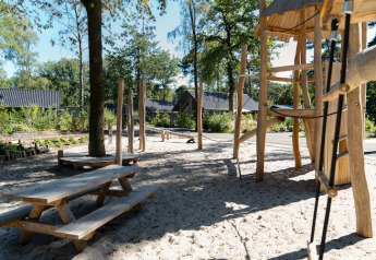 Trælegeplads med sand, borde og bænke i et udendørsområde ved en lodge omgivet af natur og hytter.