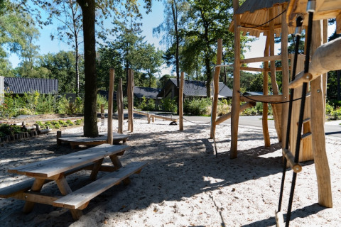 Trælegeplads med sand, borde og bænke i et udendørsområde ved en lodge omgivet af natur og hytter.