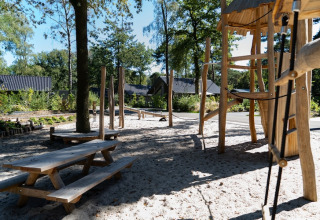 Wooden playground structure with picnic tables on sand, near a lodge surrounded by trees and cabins.