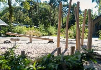 En udendørs legeplads ved en lodge med en vippe, træstolper og natur omgivelser i baggrunden.