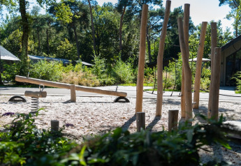 En udendørs legeplads ved en lodge med en vippe, træstolper og natur omgivelser i baggrunden.