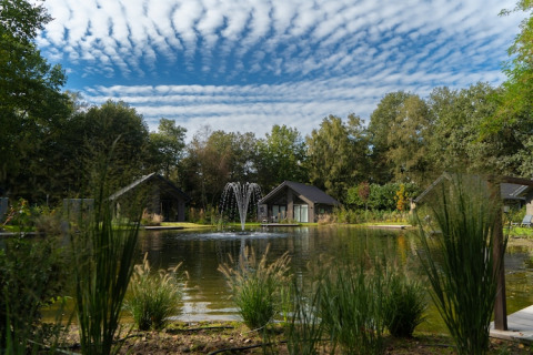 Beaux lodges près d’un étang avec une fontaine, entourés d’arbres et de ciel nuageux impressionnant.