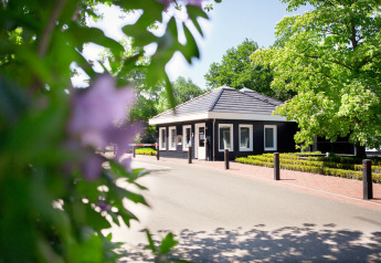 Indgang til en lodge med sorte vægge og hvide karme omgivet af grønne træer og blomster i forgrunden.