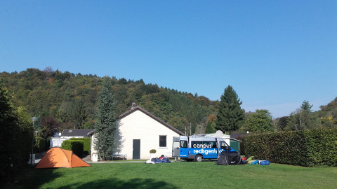Tentenkamp en busje op groene grasveld bij Camping Village Sy, met huisje, bomen en heuvels op de achtergrond.