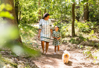 A mother and daughter walk their small dog along a woodland path near a lodge on a sunny day.