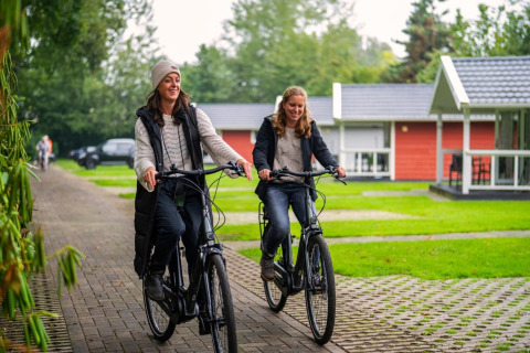 Two women happily ride bikes on a paved path in front of modern lodges at a green holiday park.