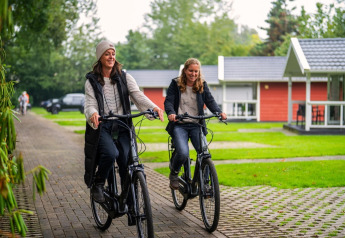 Zwei Frauen fahren lachend mit dem Fahrrad auf einem Weg vor modernen Lodges in einer grünen Anlage.