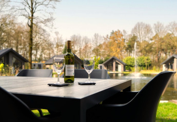 Et udendørs bord med en flaske vin, to vinglas og udsigt over søen ved moderne lodgehytter i naturen.