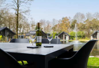 Ein Outdoor-Tisch mit Weinflasche und zwei Gläsern, Blick auf See und moderne Lodges in grüner Umgebung.