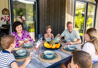 En familie nyder et måltid sammen udendørs ved et lodge, med smil og frugt i midten af bordet.
