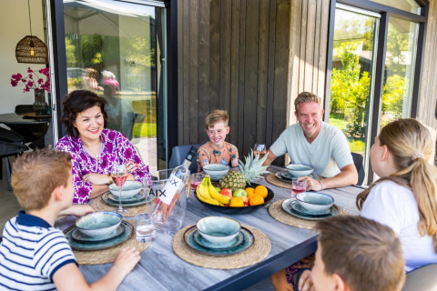 A family enjoys an outdoor meal together at a lodge, smiling around a table with fruit in the center.