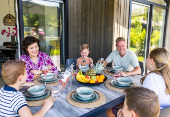 En familie nyder et måltid sammen udendørs ved et lodge, med smil og frugt i midten af bordet.