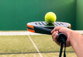 Nærbillede af en hånd, der holder et padelbat med en gul bold ovenpå, på en udendørs bane.