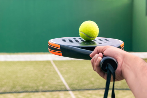Nærbillede af en hånd, der holder et padelbat med en gul bold ovenpå, på en udendørs bane.