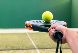 Close-up of a hand holding a padel racket with a yellow ball on top, on an outdoor court.