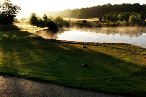 Serene morning view of a glamping site with a lush lawn, trees, and mist rising over a peaceful pond.