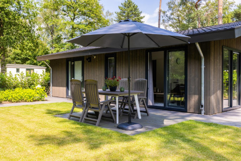 Terraza soleada en Holiday Park Latour 8, Países Bajos, con mesa de jardín, sombrilla y lodge moderno entre árboles.
