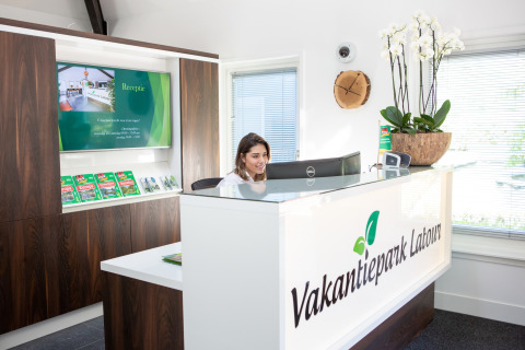 Réceptionniste au comptoir de Vakantiepark Latour, brochures, plantes et intérieur clair et moderne en arrière-plan.