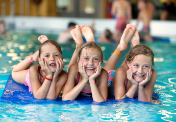 Trois enfants souriants allongés sur un tapis bleu dans la piscine d’un hébergement glamping, amusés.