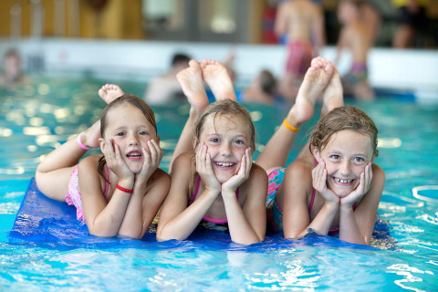 Tres niños sonrientes tumbados sobre una colchoneta azul en la piscina de un glamping, disfrutando juntos.