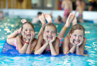 Tre glade børn leger på en blå måtte i en indendørs pool på et glamping sted, omgivet af vand.