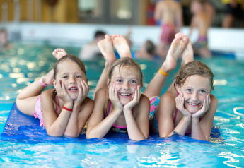 Tre glade børn leger på en blå måtte i en indendørs pool på et glamping sted, omgivet af vand.