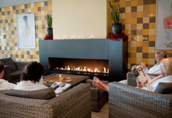 People relaxing in robes by a cozy fireplace lounge at a glamping accommodation with woven chairs.