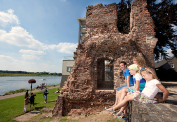 Familia sentada en ruinas junto a un río soleado, personas pasean cerca de alojamiento glamping.