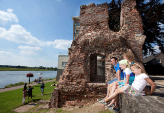 Familia sentada en ruinas junto a un río soleado, personas pasean cerca de alojamiento glamping.