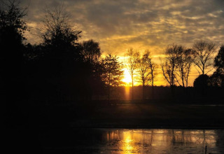 Solnedgang over et roligt naturspot med silhuetter af træer og refleksioner i vandet ved et glampingsted.