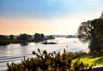 View of a serene river, trees, and a ferry boat, captured from a glamping accommodation on a sunny day.