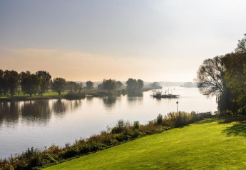 Peaceful river view with green fields, trees, and a ferry in the distance at a glamping accommodation.